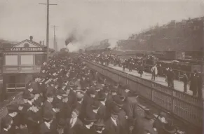 Photograph of a large group of men in hats waiting at a train station in East Pittsburg