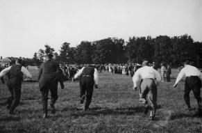 Black and white photo of men running, captured from behind, crowd in distance.