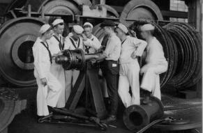 Black and white image of cadets dressed in nautical uniforms examining large machinery in a factory..