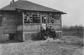Black and white image of four men in hats and suits seated on the steps of a small building in poor repair.