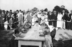 Black and white image of children lined up at a table, in front of an audience, for a pie eating contest.