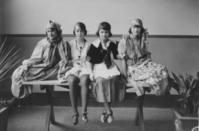 Black and white photograph of four young women in costumes, seated on a wooden table.