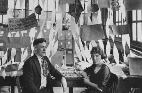 Black and white photograph of a man and woman seated in front of a desk decorated with a wedding theme.