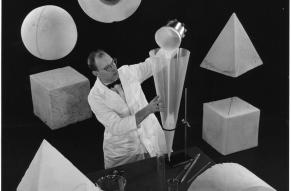 Black and white image of a scientist and scientific apparatus, surrounded by large foam and rubber shapes.