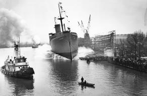 Black and white image of a large freighter ship being dropped into a busy river with many ships.