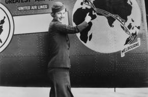 Black and white photograph of a woman in a stewardess uniform standing in front of a decorated airplane body. Plane body shows a globe with directional markers running from London to Melbourne.