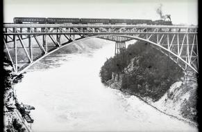 Black and white image of a train traveling on a steel arch bridge over the Niagara River rapids. 