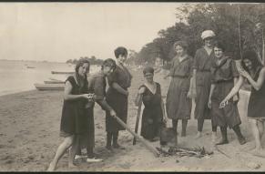 Panoramic black and white photograph of a group of women on a beach assembling a bonfire.