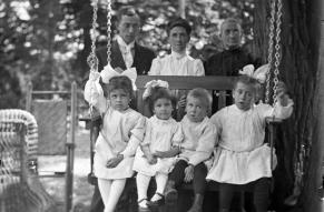 Black and white glass negative image of four children on an outdoor swing seat, with three adults behind them. 