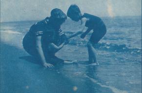 Cyanotype image of a person with two small children playing at the edge of the ocean.