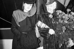 Posed portrait of two well-dressed women attending a ship launching. One is holding a ceremonial bottle and bouquet.