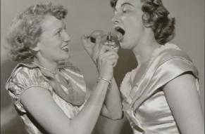 Black and white photo of two women. One is spraying something from a bottle into the other's mouth.