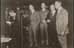 Black and white photograph of a musical pentet in a 1930s music studio. A can-shaped recording device has been emphasized with overdrawing.