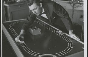 Black and white image of a man moving a model car along an electronic track