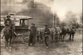 Black and white image of three men standing in front of a building and a horse-drawn wagon with 'Du Pont Powder' written on the side.