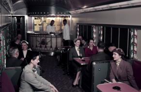 Color image of the interior of a dining car of a train, with passengers and staff.