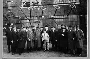 Black and white group photograph of men in suits and hats in front of an industrial building