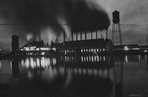 Dramatically lit black and white exterior image of a pine mill in operation at night.