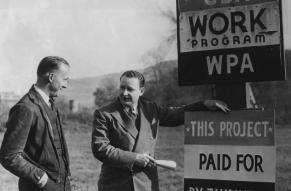 Two men standing near roadside signs declaring that roadwork is a WPA program paid for by taxes.