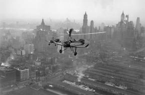 Black and white image of two autogyros flying over a cityscape.