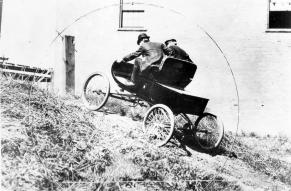 Black and white photograph of two men in a rudimentary automobile driving up a hill.