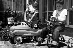 Black and white photograph of two children on a bicycle and a toy car play-acting a traffic stop.