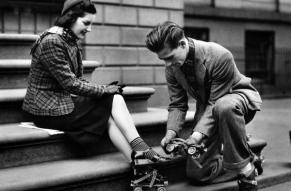 Black and white photograph of a man helping a woman strap roller skates onto her shoes.