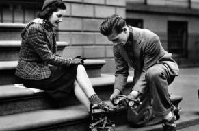 Black and white photograph of a man helping a woman strap roller skates onto her shoes.