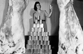 Black and white photograph of a woman with  is shown with two sides of beef and a pile of one pound tins that are equal in meat content.