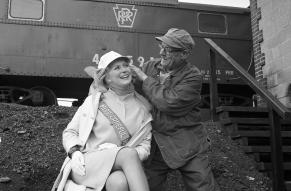 Seated woman in a sash is being crowne with a railroad workers' hat. Train in background.