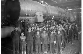 Group photograph of a large number of men in work clothes posed in front of a steam engine boiler