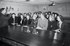 Black and white photograph of a man demonstrating protective goggles and shoes to a classroom of women.