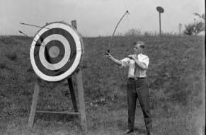 Black and white photograph of a man with a bow and arrow, posed next to a large bullseye target