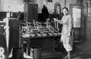 Black and white image of a woman in work clothes standing by a bolt threading machine in a workshop.
