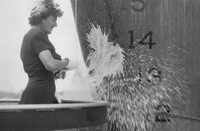 Black and white photograph of a woman christening the hull of a large ship