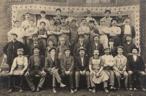 Black and white group portrait of automobile industry workers posed in front of a stars and stripped patterned banner backdrop 