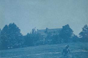 Cyanotype photo of a person sitting in a grassy field with trees and a large house in the background.