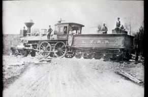 Black and white image of a ca. 1850s wood-burning locomotive, with men posed on and around it.