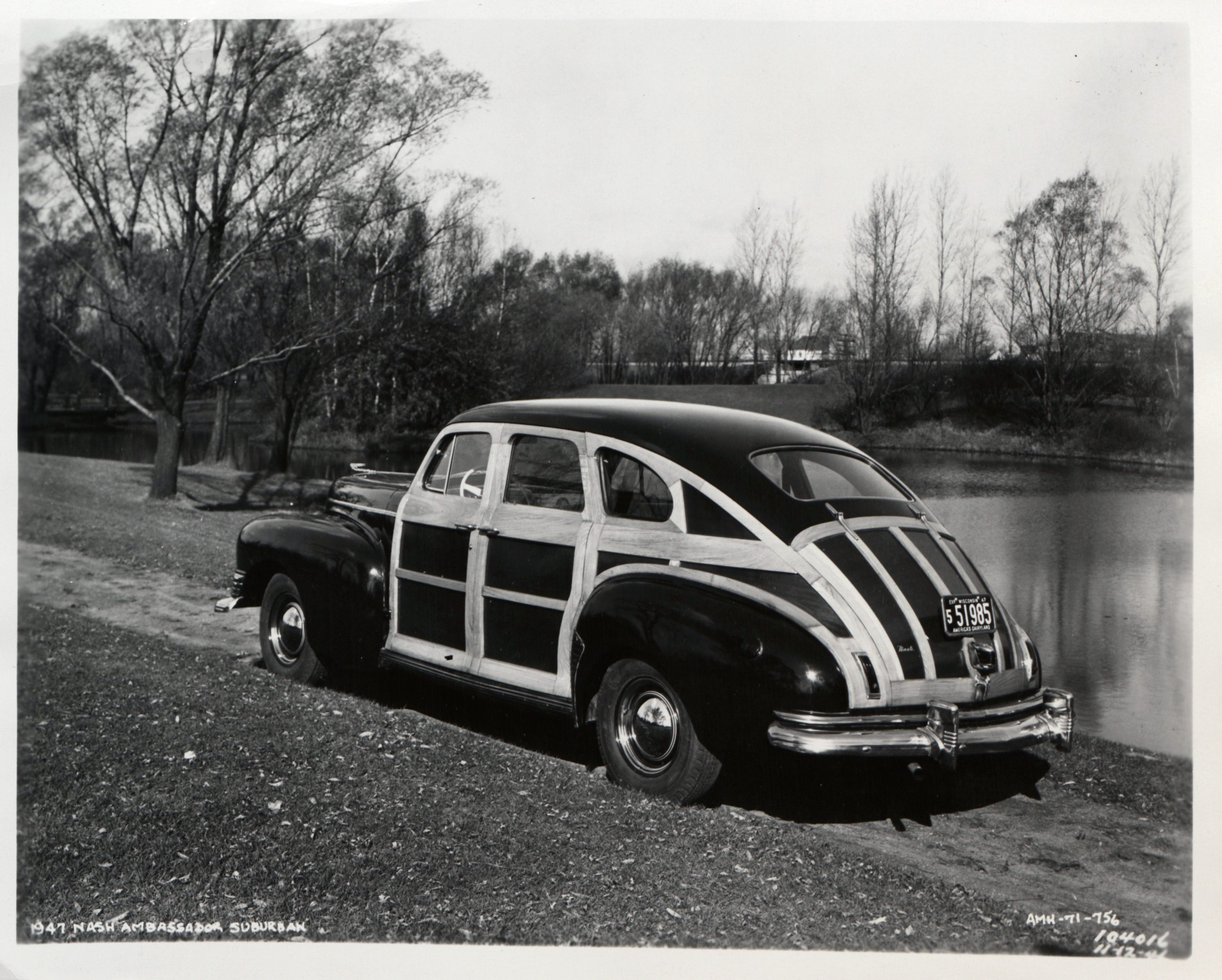 Black and white photograph of a wood-paneled 1947 Nash Ambassador Suburban parked on a dirt roadway by a body of water.